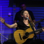 Heatherlyn sings into a microphone with a hand raised to the side and her eyes closed whilst holding an acoustic guitar. Tony Bean plays a five string bass guitar in the background.