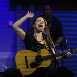 Heatherlyn sings into a microphone with a hand raised and her eyes closed whilst holding an acoustic guitar. Tony Bean plays a five string bass guitar in the background.