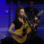 Heatherlyn sings into a microphone whilst playing an acoustic guitar. Tony Bean plays a five string bass guitar in the background.