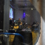 Through a small oval window the photo looks in at the Nine Beats Collective from their right hand side. In the foreground is the merch stall with the band and the front row of the audience both in view.