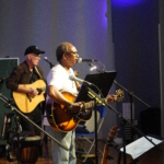 Ben Okafor performs a song whilst playing guitar. Steve Bassett plays guitar in the background and another guitar on a stand sits in the foreground.