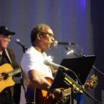 In the stage light, Ben Okafor performs a song whilst playing guitar. Steve Bassett plays guitar in the background.