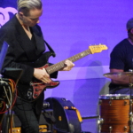 Rich Wilson plays guitar whilst Marcus Copeland sits to the right playing the drums.