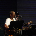 Ben Okafor performs a song whilst playing guitar. Steve Bassett plays guitar in the background.