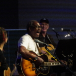 Ben Okafor performs a song whilst playing guitar. Steve Bassett smiles and plays guitar in the background and Heatherlyn stands to the side holding a ukelele.