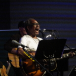 Ben Okafor performs a song whilst playing guitar. Steve Bassett plays guitar in the background and Heatherlyn stands to the side holding a ukelele.