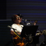 Ben Okafor performs a song whilst playing guitar. Steve Bassett plays guitar in the background.