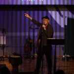 Steve Bassett performs spoken word, holding a microphone in his left hand and with his right hand raised and pointing up to the right. Tony Bean stands to the left holding a bass guitar.