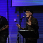 Steve Bassett performs spoken word with his hands held palms open in front of him. Tony Bean stands to the left holding a bass guitar.