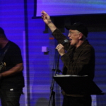 Steve Bassett performs spoken word with a hand in the air. Tony Bean stands to the left holding a bass guitar.