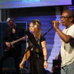 Ben Okafor sings whilst Heatherlyn stands in the background and to the left Rich Wilson plays guitar.