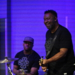 Tony Bean smiles whilst playing bass guitar. Marcus Copeland plays drums in the background.