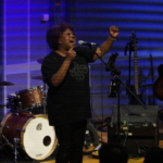 Belinda Kae sings passionately into a microphone with her left arm raised. Heatherlyn sits with a ukulele to the side.