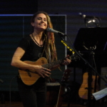 Heatherlyn plays a ukulele and smiles at the audience whilst stood in front of a microphone.