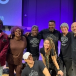 Ben Okafor, Belinda Kae, Marcus Copeland, Tony Bean, Steve Bassett and photographer Jason stand posed for the photo. Martin Trotman and Heatherlyn pose in front. The wall behind them is lit up purple.